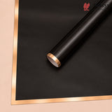A roll of matte black wrapping paper or vinyl with a metallic rose gold edge, shown diagonally on a beige background with a matching flat sheet of the same material.