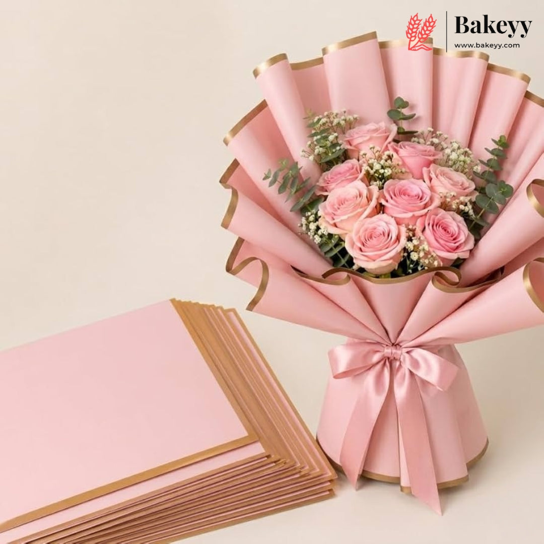 A pink floral bouquet wrapped in pink paper with gold edges and tied with a satin ribbon, shown beside a stack of matching pink wrapping sheets on a light beige background. The Bakeyy logo appears in the top right corner.