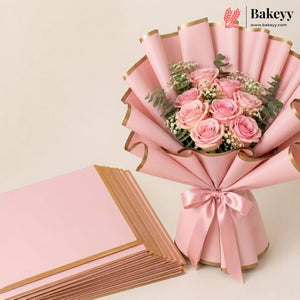 A pink floral bouquet wrapped in pink paper with gold edges and tied with a satin ribbon, shown beside a stack of matching pink wrapping sheets on a light beige background. The Bakeyy logo appears in the top right corner.