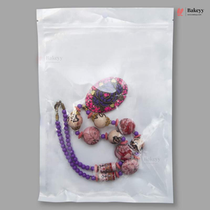 A clear plastic zip-top bag containing a decorative beaded necklace or bracelet with purple beads, pink and white patterned round beads, and a large oval floral pendant, shown on a gray background. The Bakeyy logo appears in the top right corner.