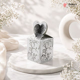 Silver decorative box with heart-shaped lid on a marble surface with white flowers.