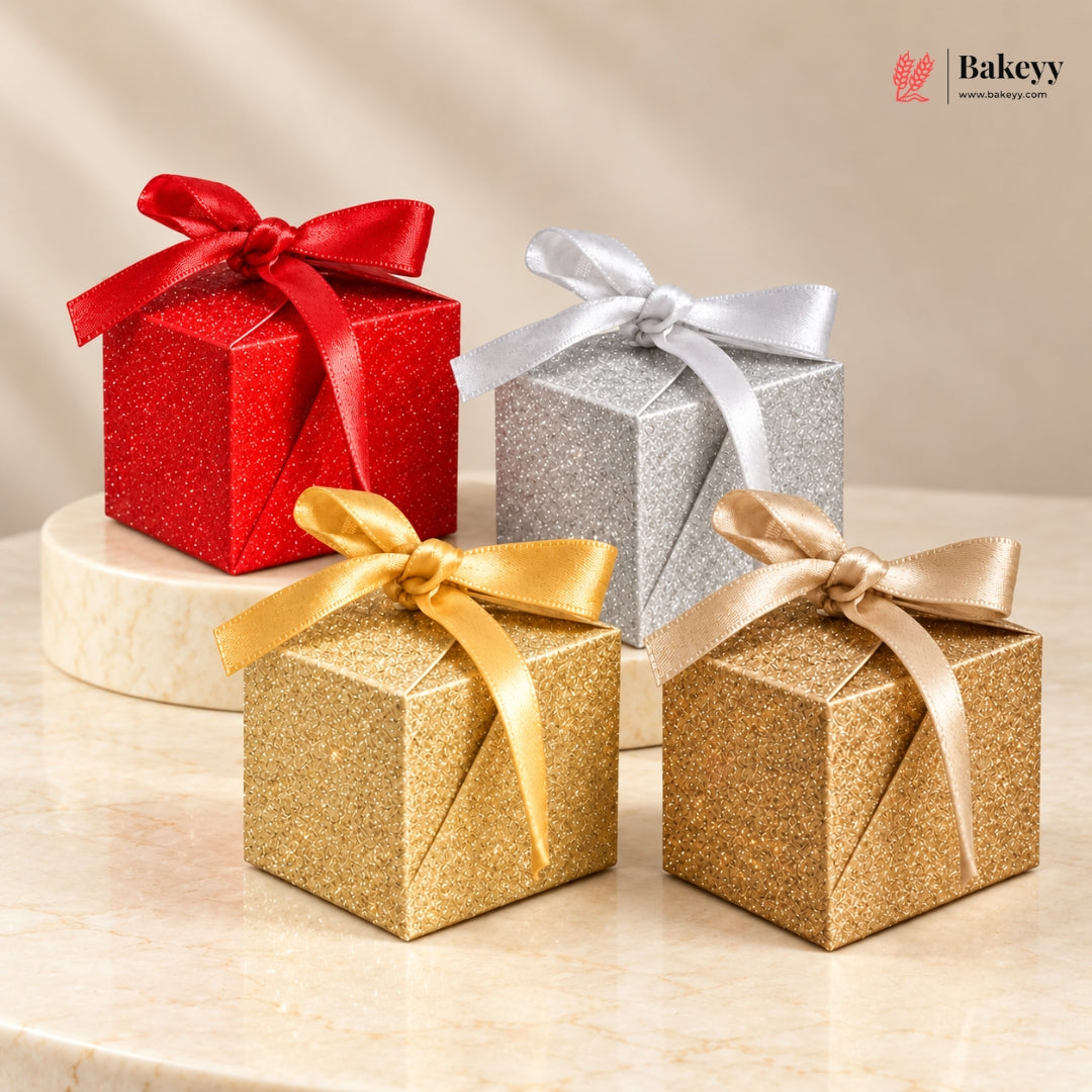 Four glittery cube-shaped gift boxes in red, silver, and gold, each tied with a satin ribbon, arranged on a light marble surface with a soft beige background. A Bakeyy logo appears in the top right corner.
