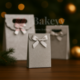 Silver gift bag with a bow on a wooden surface with blurred lights in the background, featuring the brand 'Bakeyy'.