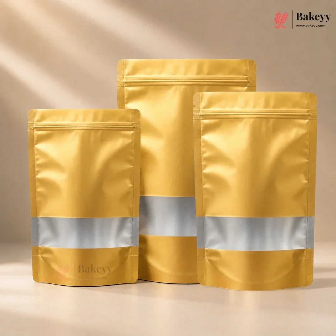 Three metallic gold stand-up pouches with reflective silver bands, arranged in a studio setting against a warm beige background. The Bakeyy brand logo is visible in the top right corner and subtly printed on the bottom of the pouches.