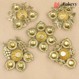 Five ornate gold-plated brass wall decor pieces with traditional Indian motifs like peacocks, grapes, and floral patterns, arranged on a solid beige background. A 'Bakeyy' brand logo is visible in the top right corner.