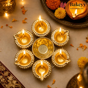 A decorative arrangement of six gold-toned oil lamps (diyas) with lit flames, centered on a neutral surface with warm ambient lighting and festive flower petals. A Bakeyy logo watermark is visible in the top right corner.
