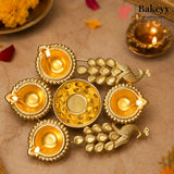 A decorative brass Diwali oil lamp set featuring five small lamp holders arranged around a central round lamp, with peacock motifs between them. The lamps are lit and displayed on a beige surface with floral props and a Bakeyy brand watermark in the top right.