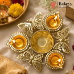 A brass diya holder with five butterfly-shaped candle holders arranged around a central bowl, lit with five burning candles, on a beige fabric background with decorative flowers and a tray in the background. The Bakeyy logo is visible in the top right corner.