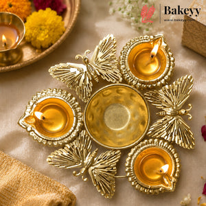 A brass diya holder with five butterfly-shaped candle holders arranged around a central bowl, lit with five burning candles, on a beige fabric background with decorative flowers and a tray in the background. The Bakeyy logo is visible in the top right corner.