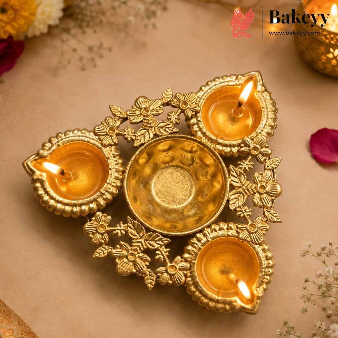 A decorative brass four-light diya holder with three lit candles and one central candle cup, styled on a beige surface with scattered flower petals and a Bakeyy logo in the top right corner.