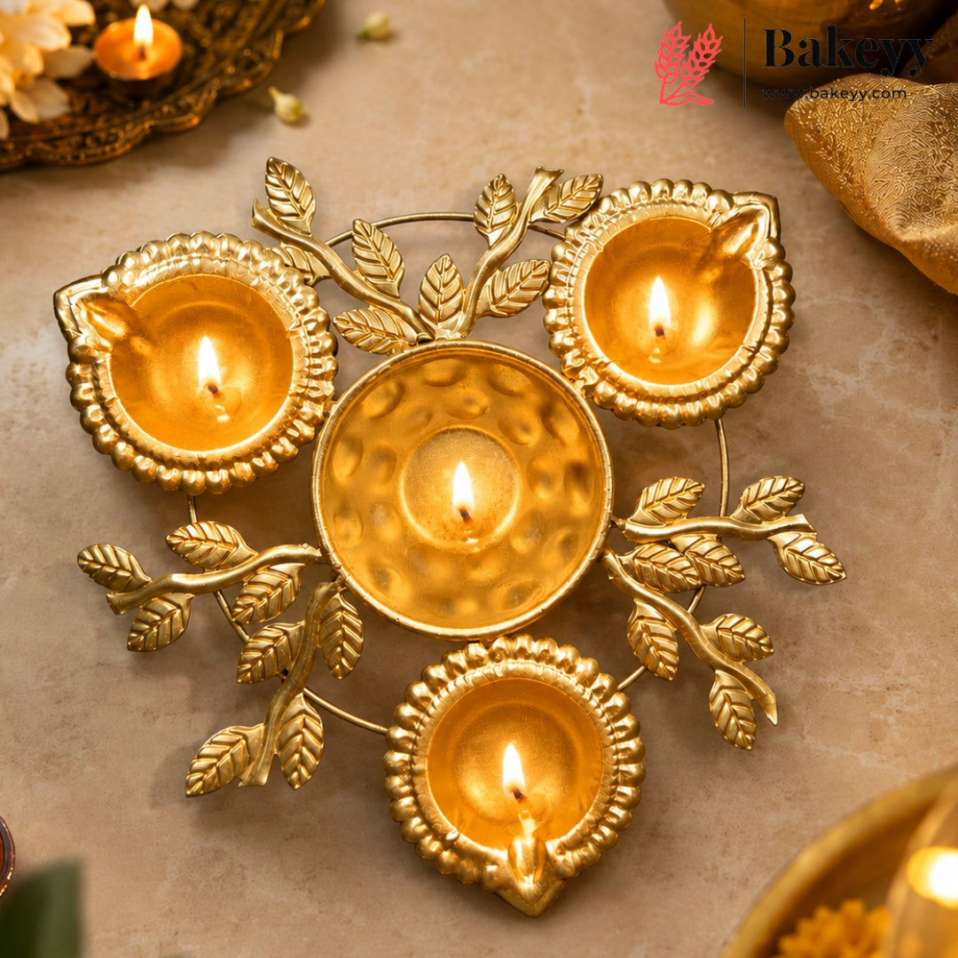 A decorative gold-toned metal diya holder with four lit tealight candles, styled on a beige stone surface with warm ambient lighting and a Bakezy logo watermark in the top right.