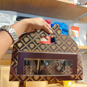 A hand wearing a silver watch holds a decorative brown and gold patterned bakery box with a clear window displaying marbled pastries inside, set on a wooden shelf with packaged goods in the background.
