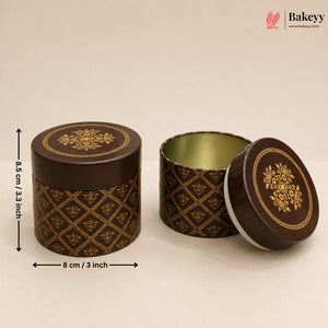 Two round decorative metal gift boxes with lids, shown on a plain beige background. One box is closed and standing upright, showing a dark brown exterior with a gold floral and geometric pattern, and a gold-toned interior.