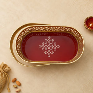 An oval gold-toned serving tray with a carved floral border and a deep red interior, decorated with a white geometric motif in the center. The tray is photographed on a beige textured surface with a small lit diya in the upper right and a burlap sack with almonds in the lower left.