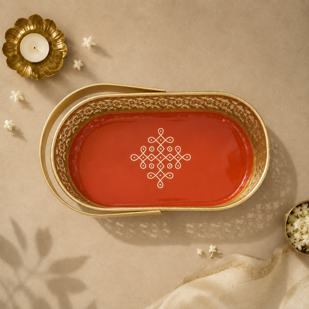 An oval decorative tray with a gold metal frame and handles, featuring a glossy red-orange interior with a white geometric line pattern in the center. The tray is photographed from above on a beige surface with a candle, small flowers, and draped fabric as styled props.