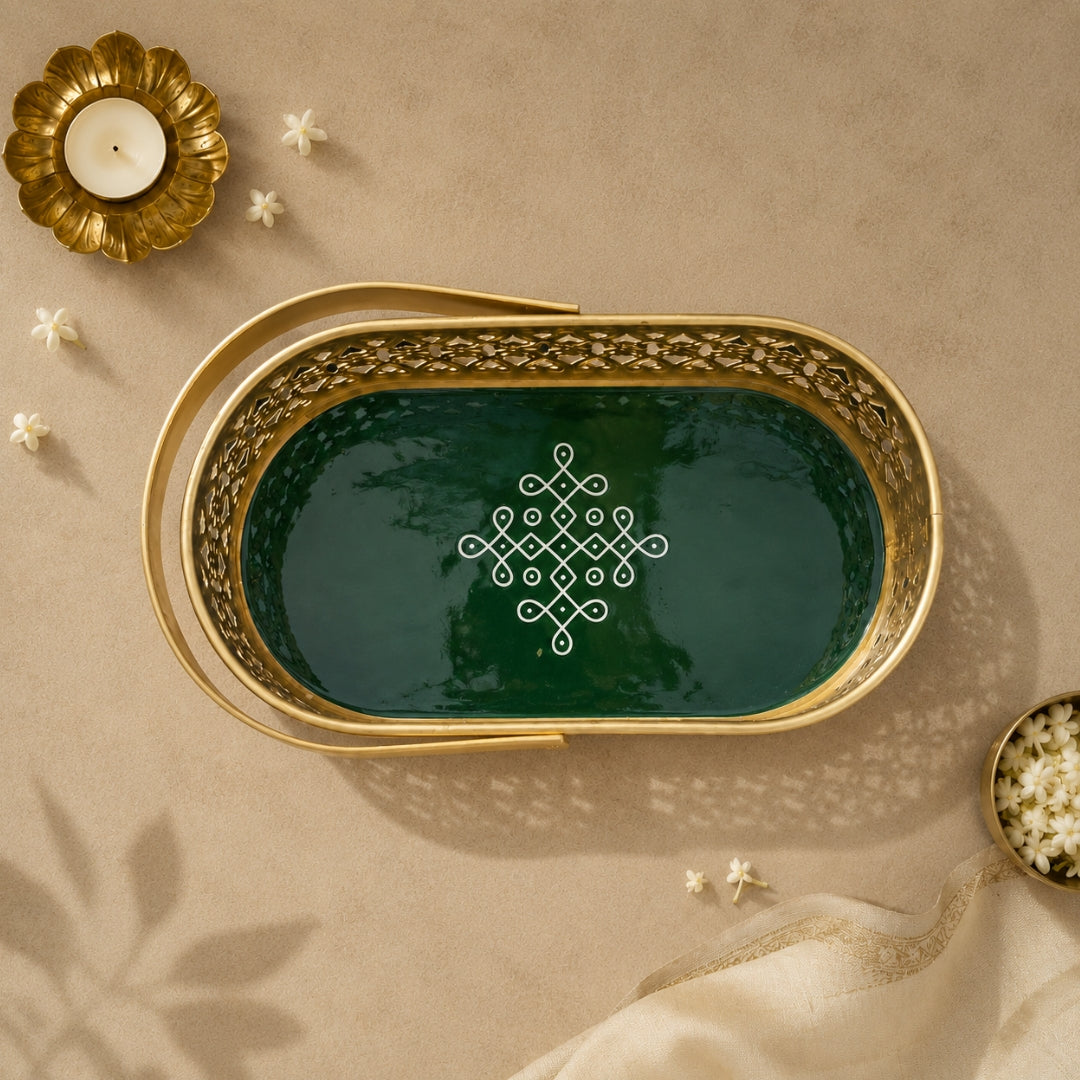 An oval decorative tray with a gold-toned metal frame and a dark green interior, featuring an intricate white geometric pattern in the center. The tray is styled in a flat-lay setting on a beige textured surface with a gold candle holder, small white flowers, and a cream fabric napkin.