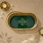 An oval decorative tray with a gold-toned metal frame and a dark green interior, featuring an intricate white geometric pattern in the center. The tray is styled in a flat-lay setting on a beige textured surface with a gold candle holder, small white flowers, and a cream fabric napkin.