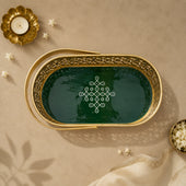 An oval decorative tray with a gold-toned metal frame and a dark green interior, featuring an intricate white geometric pattern in the center. The tray is styled in a flat-lay setting on a beige textured surface with a gold candle holder, small white flowers, and a cream fabric napkin.