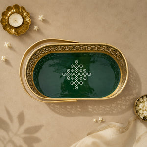 An oval decorative tray with a gold-toned metal frame and a dark green interior, featuring an intricate white geometric pattern in the center. The tray is styled in a flat-lay setting on a beige textured surface with a gold candle holder, small white flowers, and a cream fabric napkin.