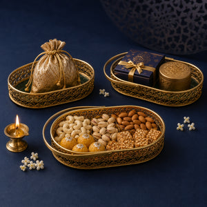 Three ornate gold trays arranged on a dark navy surface, filled with assorted sweets, nuts, and a small wrapped gift box. A brass diya lamp glows at left, with scattered white flowers and a decorative dark blue object in the background.