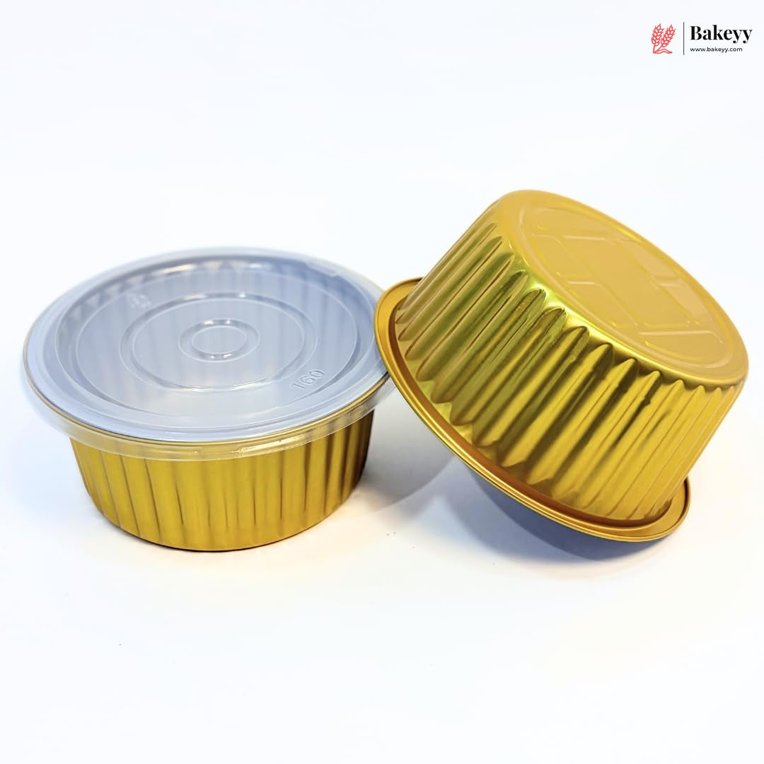 Two small gold fluted disposable food containers on a white background, one upright with a clear plastic lid and one tipped over to show the gold base.