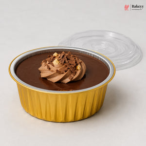 A chocolate dessert in a gold fluted disposable container with a clear lid, photographed on a light neutral background. The dessert has a swirl of chocolate frosting topped with chocolate shavings and small nut pieces.