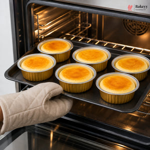 A hand wearing a beige quilted oven mitt slides a baking tray with six freshly baked mini tarts or pastries into an oven. The Bakeey logo is visible in the top right corner.