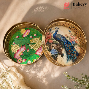 Two round gold metal trays with cutout edges, shown open on a beige surface. One tray has a green background with pink lotus flowers and gold pavements; the other has a peacock and floral scene on a cream background.