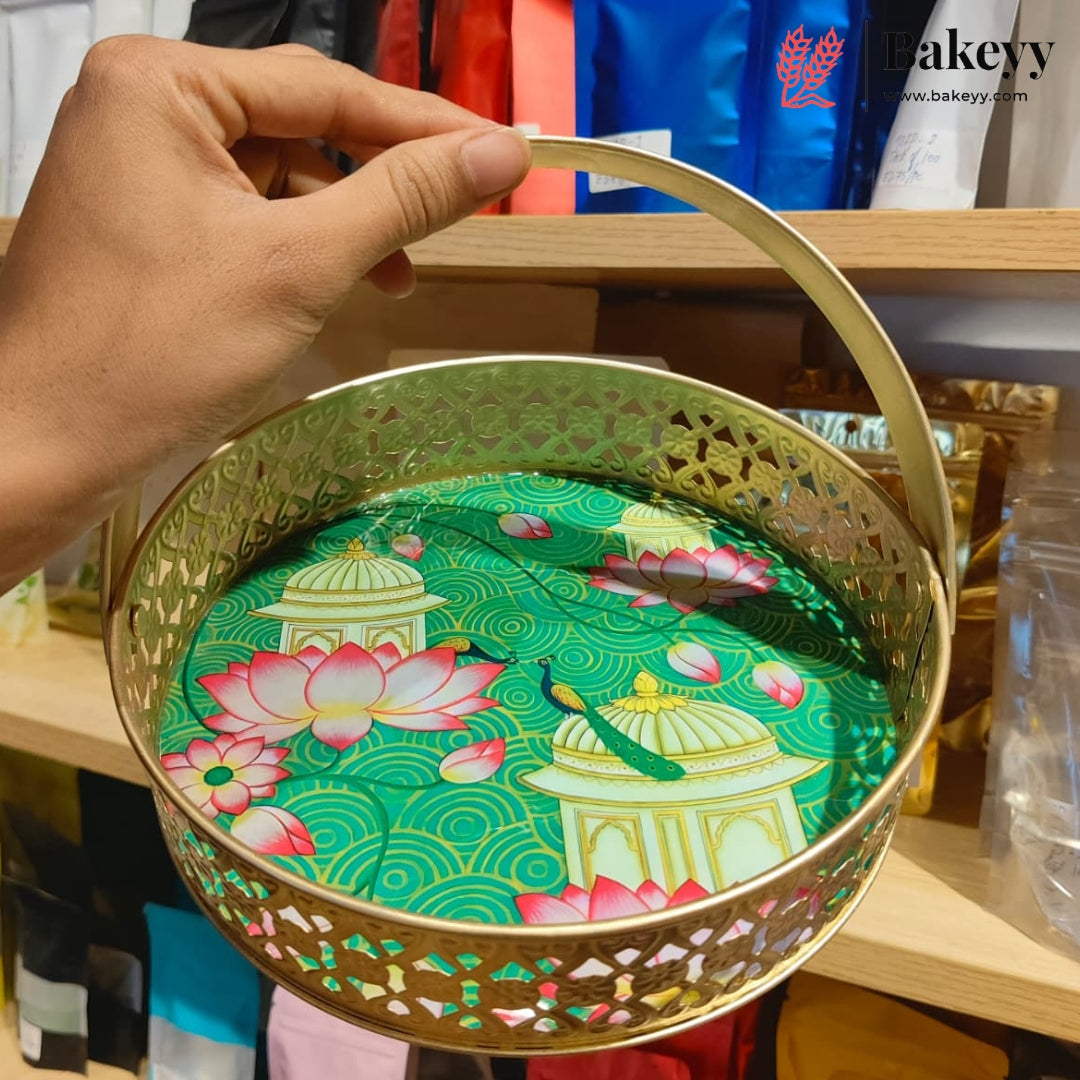 A hand holds a round gold metal basket with a handle, showing a green printed insert featuring pink lotus flowers and white domed pavilion designs. The background shows wooden shelves with packaged goods and a Bakeyy logo watermark in the top right.