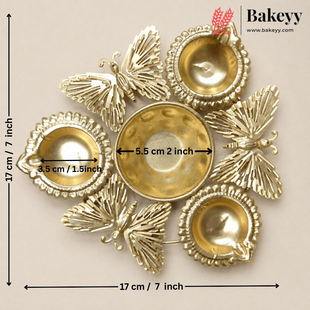 Gold-toned metal diya holder set with butterfly-shaped decorative elements and three small bowls, shown on a beige background with printed dimensions and Bakeyy branding.