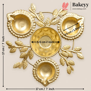 Gold-plated brass wall-mounted diya holder with three bowls and decorative leaf motifs, shown with dimension labels on a light beige background. Bakeyy logo is visible in the top right.