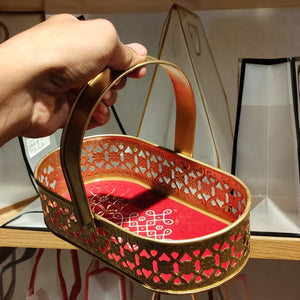 A hand holding a small oval gold-toned metal basket with a red interior and decorative cutout pattern, shown in a retail or storage setting.