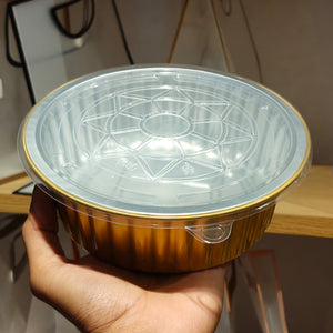 A hand holding a round disposable food container with a clear plastic lid and a metallic gold foil base, set against an indoor background.