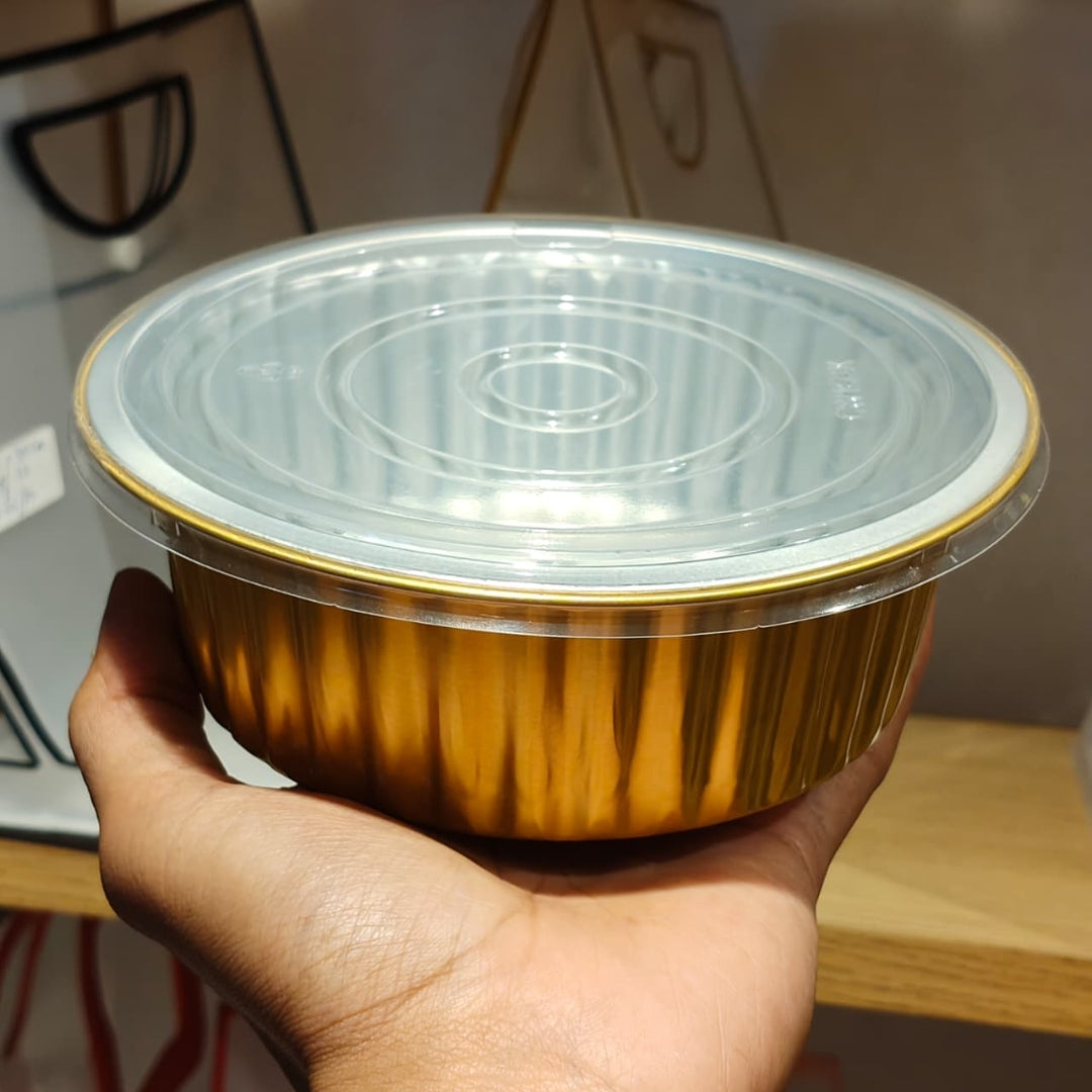 A hand holding a disposable aluminum food container with a clear plastic lid, showing the reflective gold-toned side of the container.