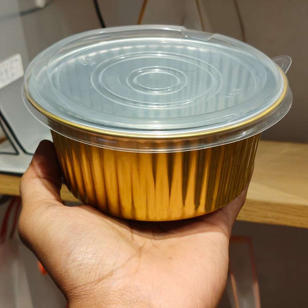 A hand holding a round disposable food container with a metallic gold foil body and a clear plastic lid, set against an indoor background.