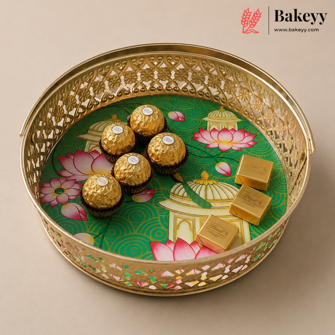 A round gold-toned decorative tray with intricate cutout detailing, holding five Ferrero Rocher chocolates and three gold-wrapped chocolate bars over a green printed insert with pink lotus motifs. The Bakeyy logo appears in the top right corner.