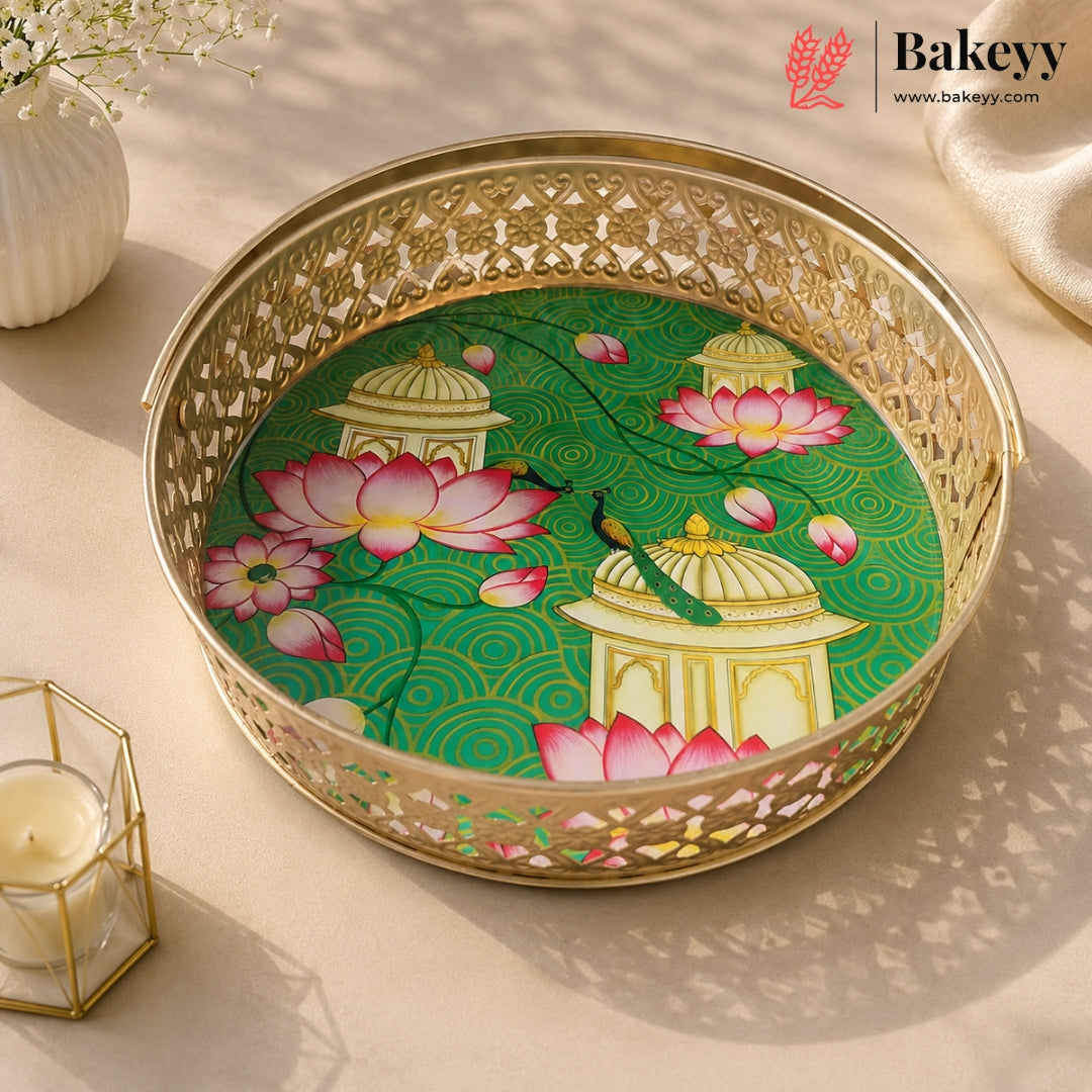 A round gold-plated metal tray with intricate cutout patterns, featuring a printed green and pink lotus pond design with domed pavements. The tray is styled on a beige surface with soft shadows, a lit candle in a geometric holder, and a white vase in the background.