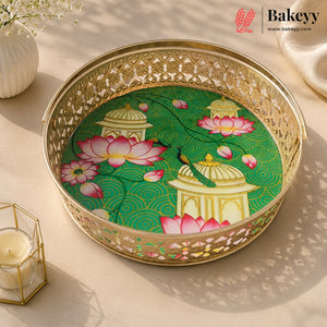 A round gold-plated metal tray with intricate cutout patterns, featuring a printed green and pink lotus pond design with domed pavements. The tray is styled on a beige surface with soft shadows, a lit candle in a geometric holder, and a white vase in the background.