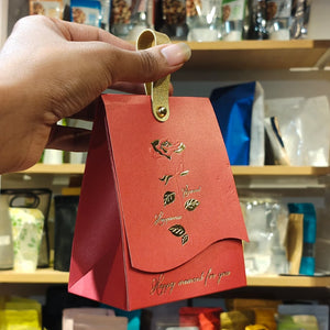 A hand holding a red paper gift bag with a gold metal clip and gold foil designs, set against a blurred background of shelves containing various packaged items.
