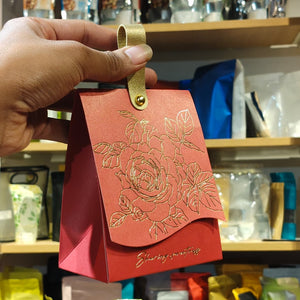 A hand holding a small red gift bag with a gold metallic strap and gold embossed floral design on the front flap, set against a blurred background of shelves with packaged items.