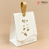 A white paper gift box with a gold hanging loop and gold foil leaf illustrations, photographed on a warm beige background. The box has the words 'Happy moment for you' printed in gold near the bottom.