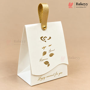A white paper gift box with a gold hanging loop and gold foil leaf illustrations, photographed on a warm beige background. The box has the words 'Happy moment for you' printed in gold near the bottom.
