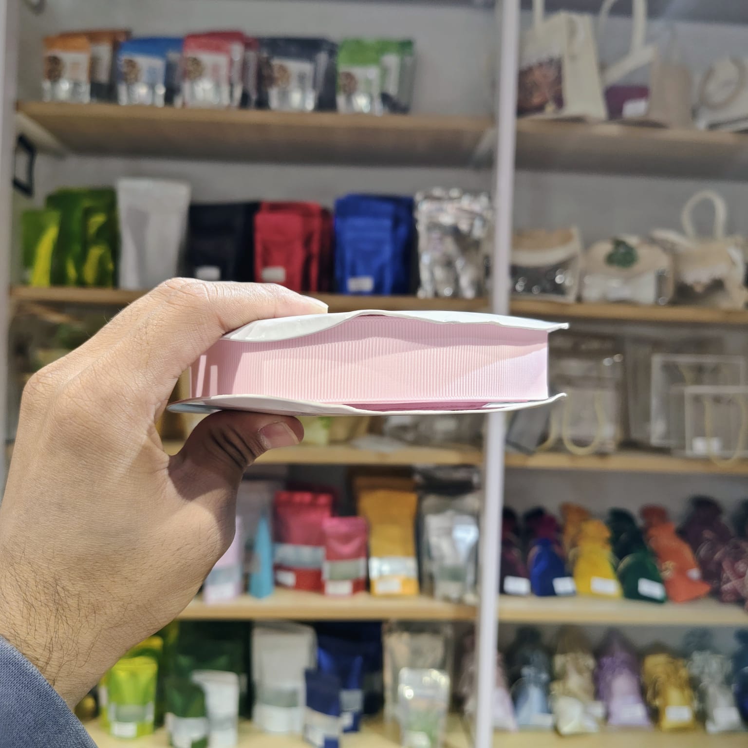 A hand holds a pink and white rectangular product, likely a book or notebook, in front of a blurred store shelf background filled with colorful packaged items.