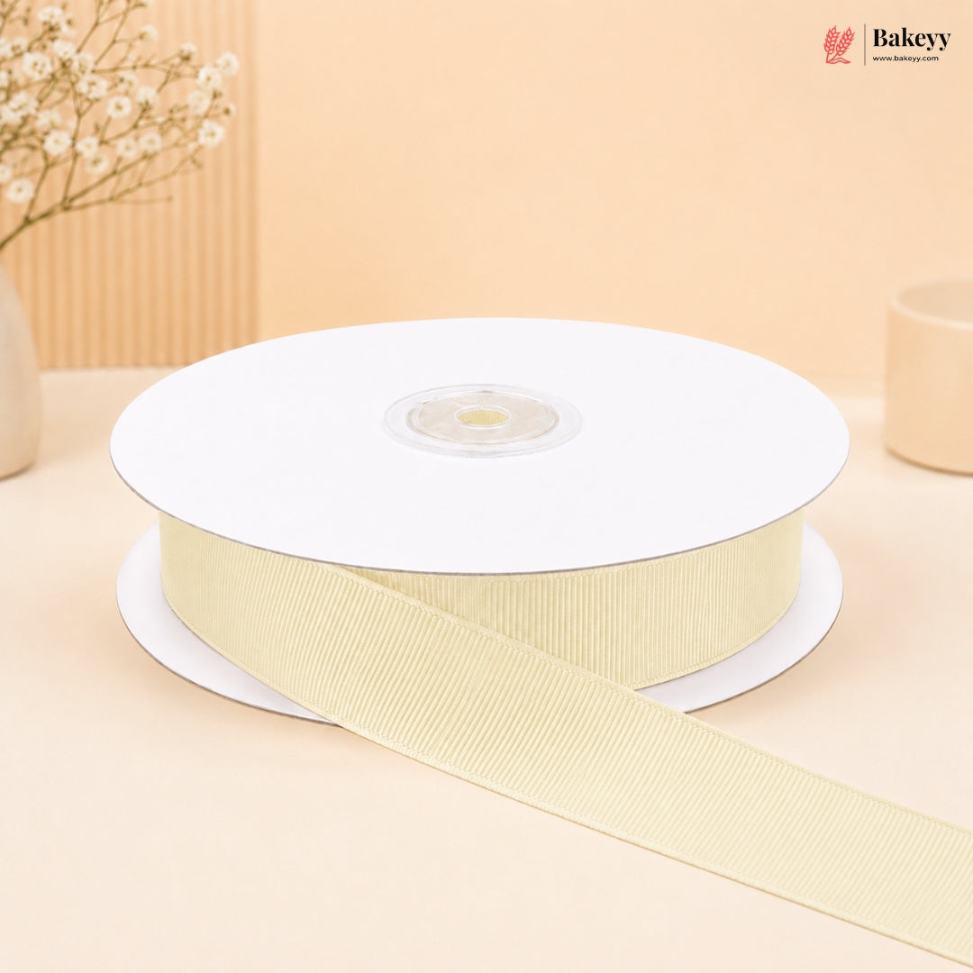 A spool of ribbed grosgrain ribbon in a pale cream color, with a length of the ribbon unspooled across a light beige surface. The background is a soft beige with a decorative dried flower vase on the left and a small container on the right.