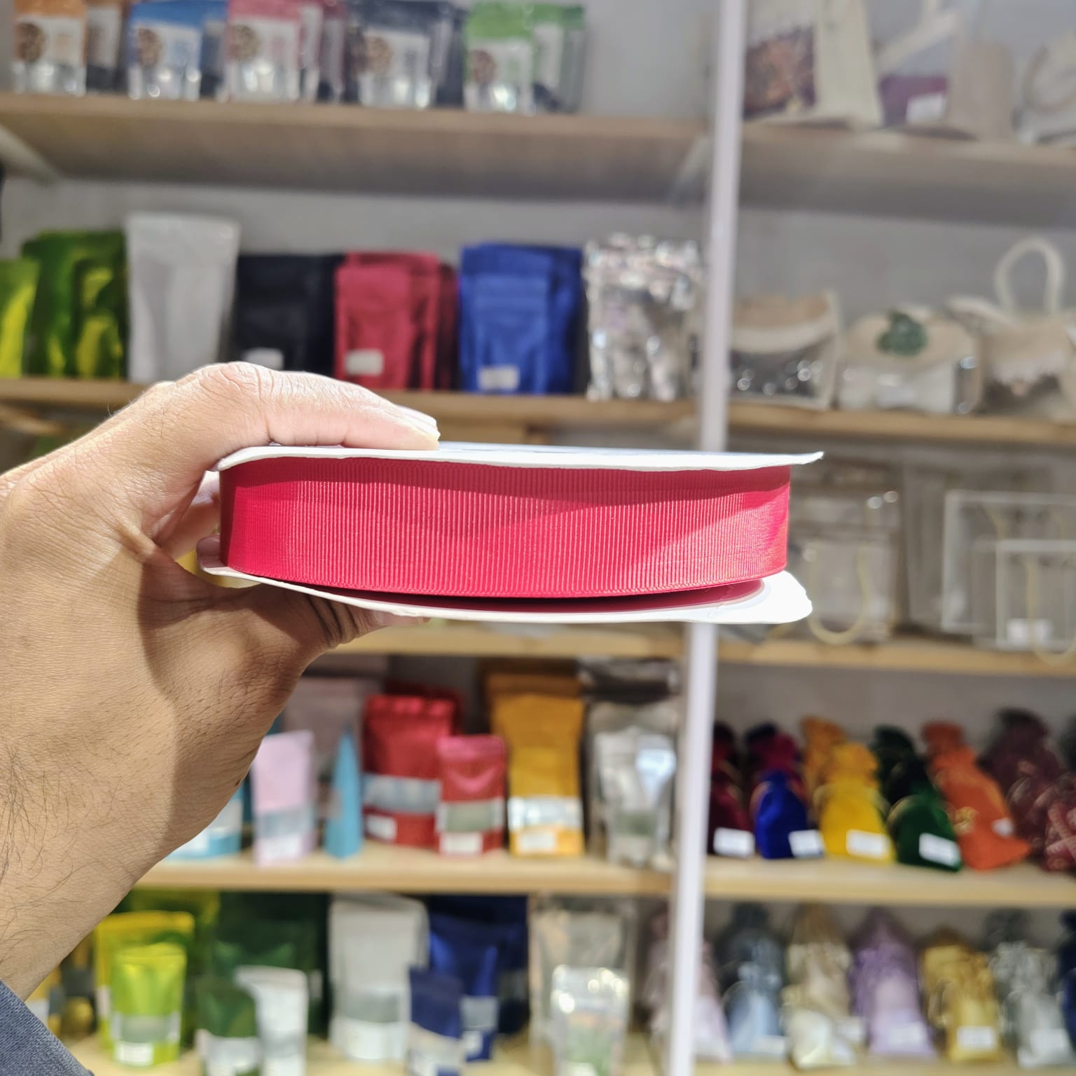 A hand holding a spool of bright pink ribbon in front of a blurred store shelf background filled with colorful packaged items.