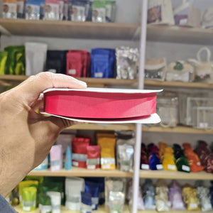 A hand holding a spool of bright pink ribbon in front of a blurred store shelf background filled with colorful packaged items.