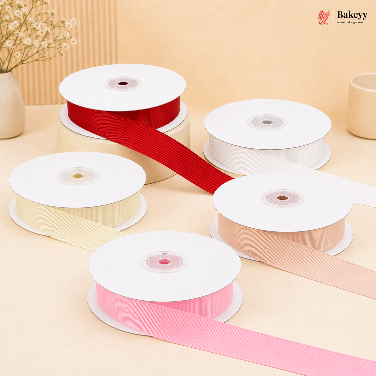 Five spools of ribbon in red, cream, white, pink, and peach, with some ribbons unspooled, arranged on a beige surface with a decorative dried flower vase in the background. The Bakeyy logo is visible in the top right corner.