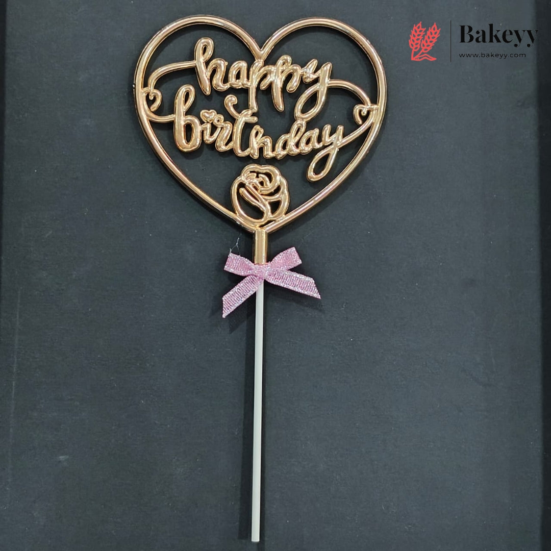 A heart-shaped cake topper with 'happy birthday' written in cursive, featuring a rose design at the center and a pink glitter bow on a stick, set against a dark gray background.