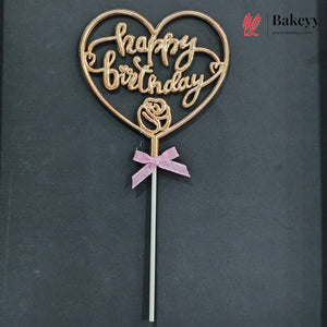A heart-shaped cake topper with 'happy birthday' written in cursive, featuring a rose design at the center and a pink glitter bow on a stick, set against a dark gray background.