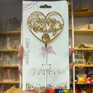 A gold heart-shaped cake topper with 'happy birthday' script and a rose design, attached to a stick with a pink glitter bow. It is displayed on its white retail packaging card, held by a hand in front of a blurred store background.